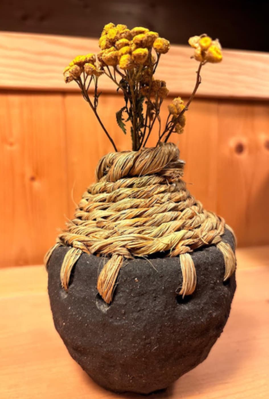 Pieza de barro oscuro con cuerda trenzada y flores secas