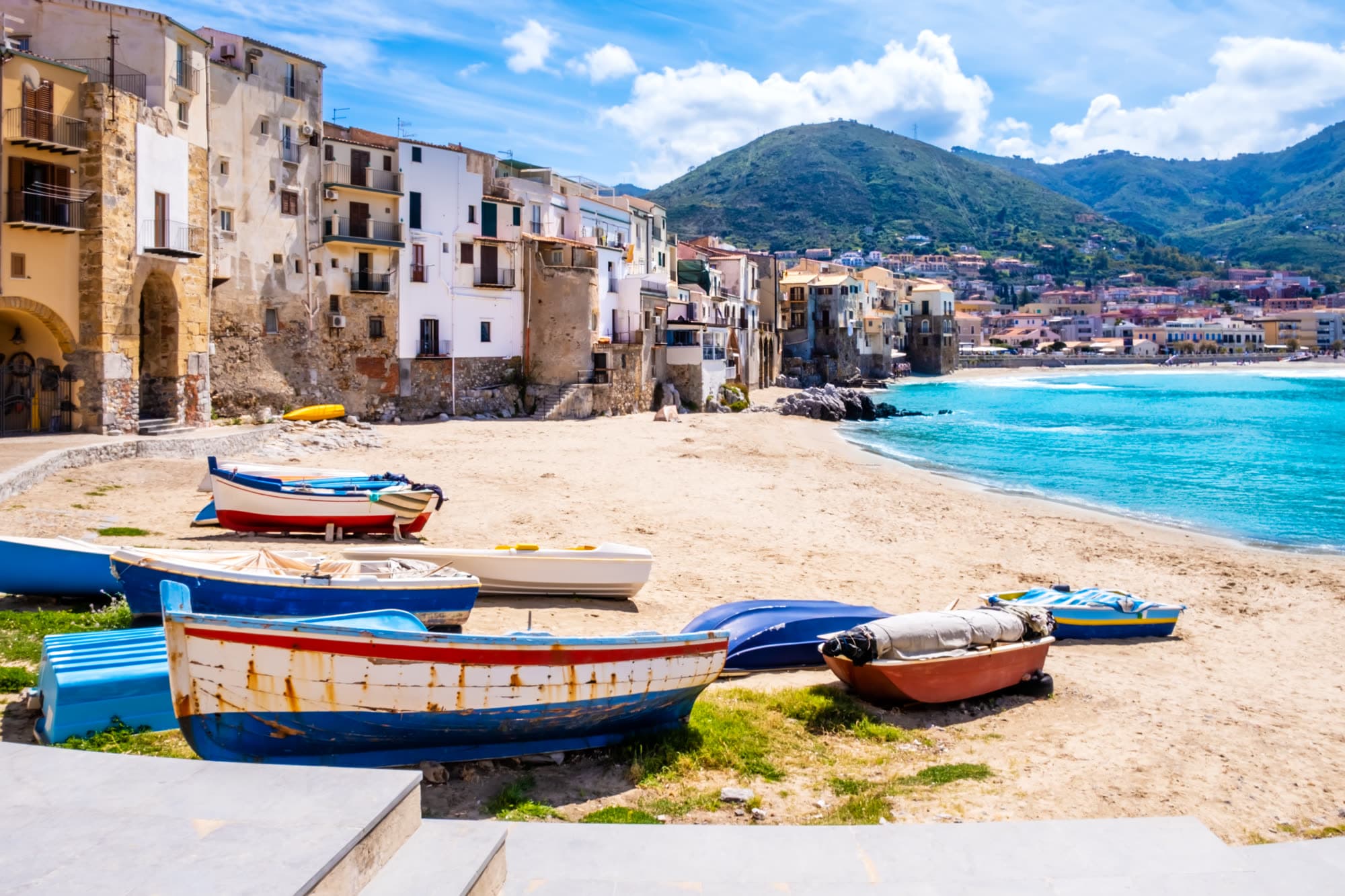 Barcas de pesca varadas en la arena de un pueblo costero mediterráneo, con casas de piedra y el mar turquesa al fondo.