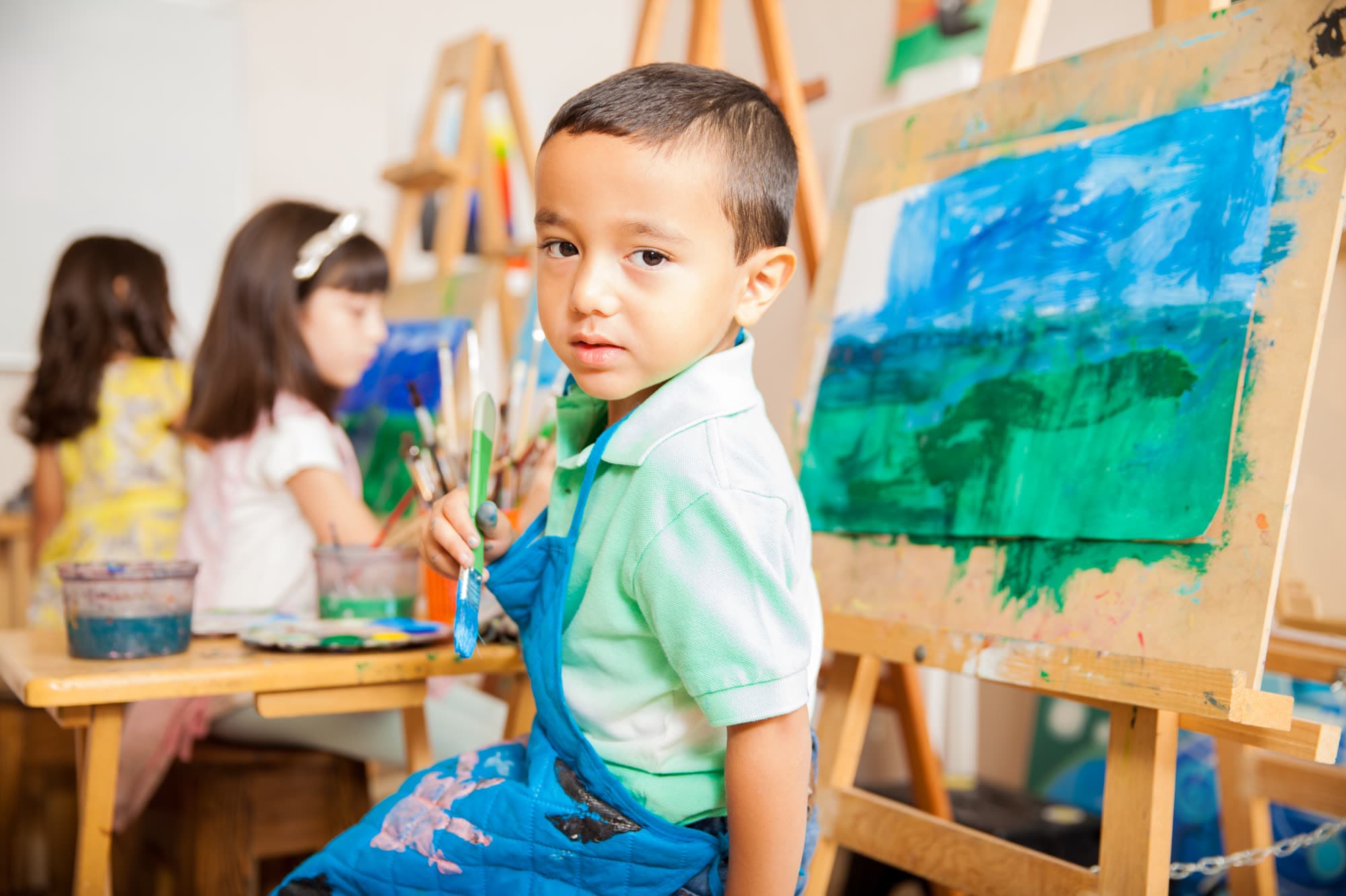 Un niño pinta con pincel un cuadro de azules y verdes en un caballete, con otras niñas pintando al fondo del taller.