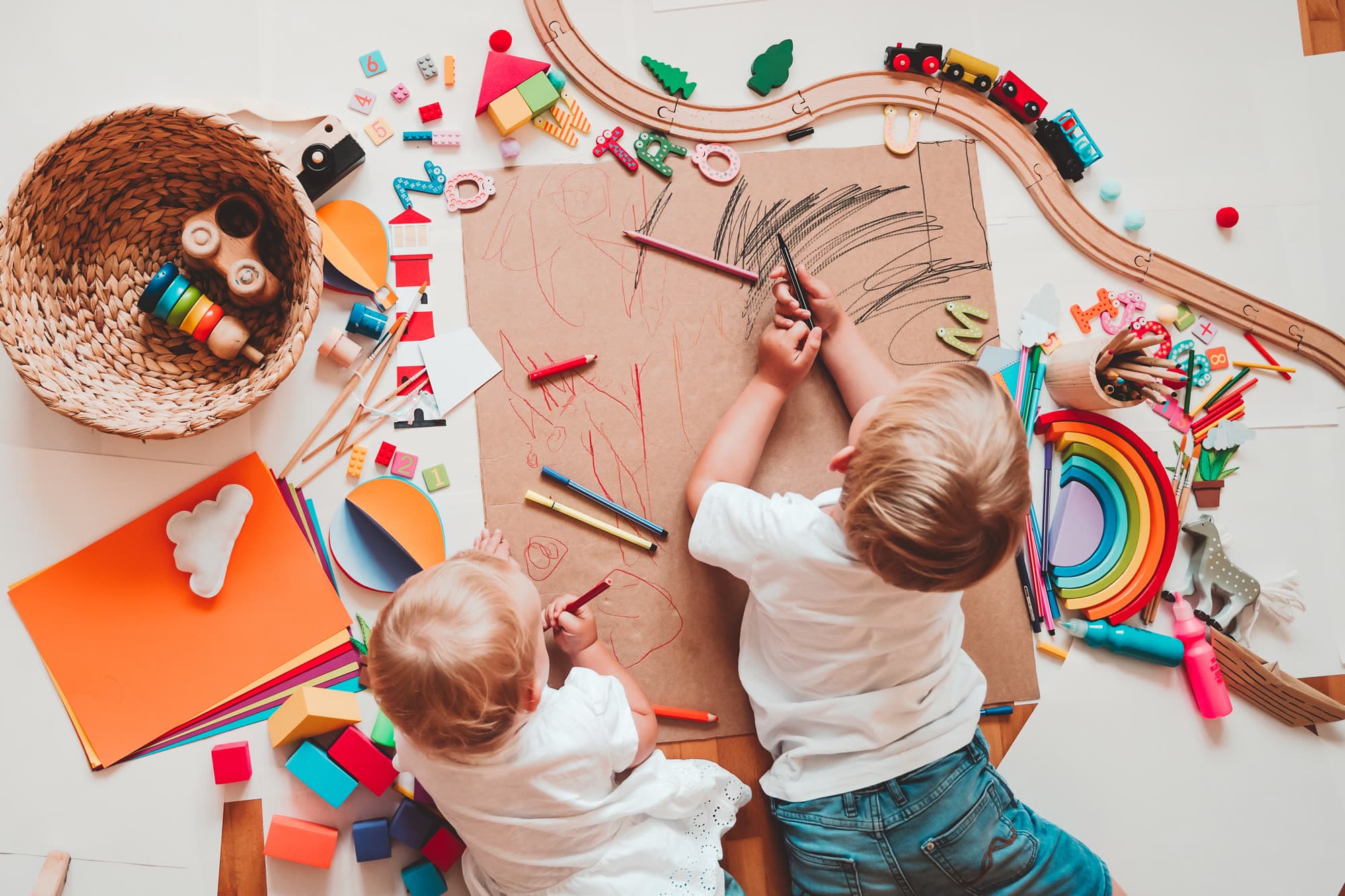 Dos niños dibujan con lápices sobre un papel grande en el suelo, rodeados de cartulinas, bloques y juguetes de madera.