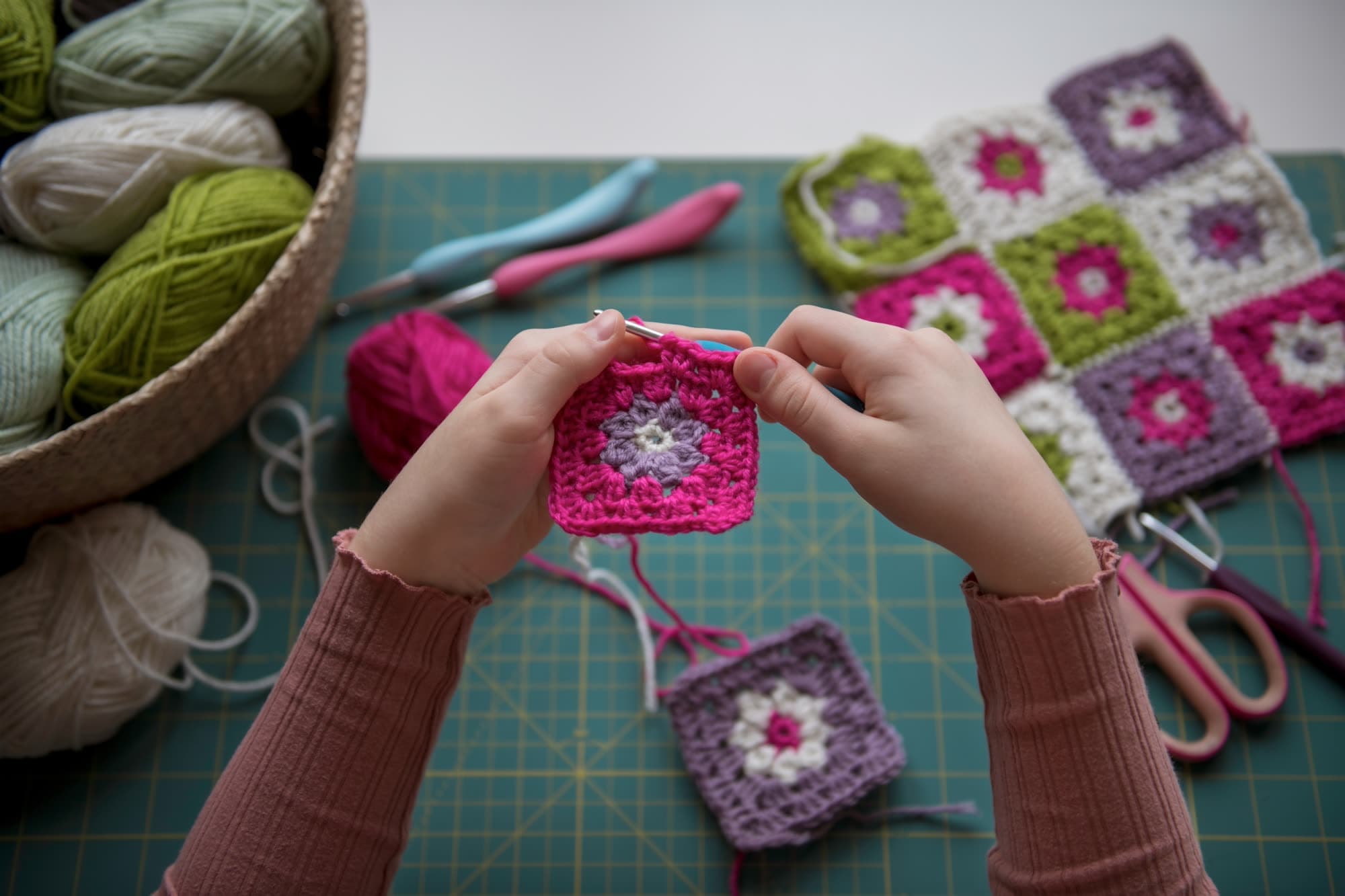 Manos tejiendo a crochet cuadrados de colores con lana y aguja.